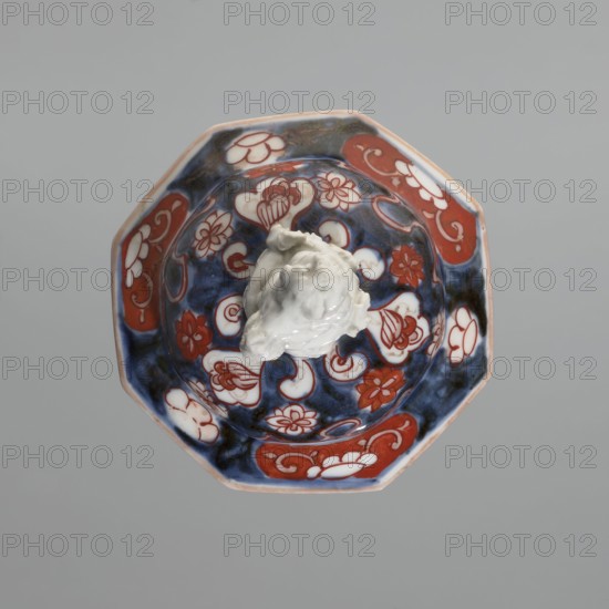 Vase with cover, Japanese, Edo period 18th century, imari ceramics, decorated with flowers, tree, bamboos, peonies, and karashishi. Vessel, height 25.5 cm, diameter 16 cm., A top-down view of an octagonal ceramic bowl with a glossy finish showing a decorative pattern in deep blue, red, white, and touches of black, containing a white irregularly shaped mound of a glossy substance; the bowl interior features floral and cloud-like motifs in red and white set against a blue ground, with a thin pale rim around the edge, all set on a plain light gray background.