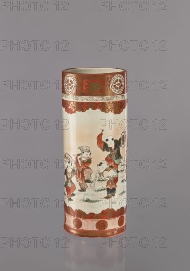 Vase, Kutani; porcelain cylinder vessel decorated with figurative scenes of one hundred sons boys and plant ornaments; ceramics; Japanese; 1868–1912; height 30 cm, diameter 12 cm., A tall cylindrical ceramic vase stands against a plain gray background, primarily showing colors of red, cream, green, black, and gold. The top rim has a decorative red band with cream and gold ornamental motifs, followed by a scalloped red and gold border. The main body features a painted scene of a group of children wearing traditional clothing in shades of red, cream, green, black, and brown, with some small green grassy accents beneath their feet. The lower section repeats a scalloped red and gold border and a horizontal red band with circular motifs, and the vase sits on a reflective surface that shows a faint mirrored image of its base.