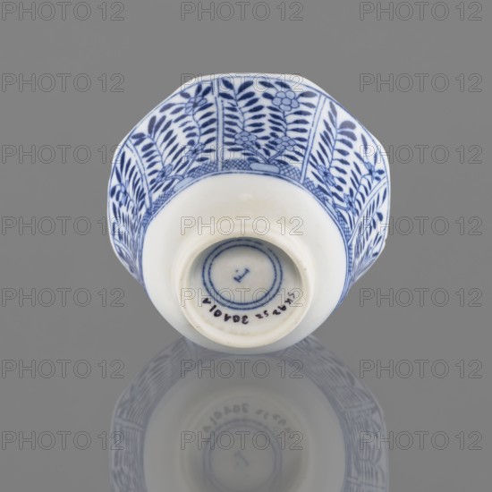 Bowl, Chinese, 20th century, ceramics vessel, height 5.3 cm, diameter 7.8 cm, A small ceramic cup is shown against a gray background, positioned rim-up so the base faces the viewer; the cup is white with a blue floral and leaf pattern around its exterior, featuring vertical panels of stylized leaves and flowers in dark blue on a white ground, and a narrow band of crosshatching near the rim. The base is recessed and plain white with a few concentric blue rings visible inside the foot, and the cup casts a soft reflection on the glossy surface beneath it. Colors visible include white, dark blue, and gray.