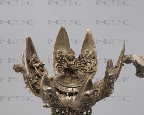 Lotus flower with Buddhist deities, 19th century, Nepal, bronze cult object in the form of a lotus mandala depicting Buddhist deities bodhisattva iconography, lotus iconography, pairs of lovers, enlightenment, height 20.3 cm, width 8.0 cm, A metallic sculptural object shaped like an open lotus with a central figure standing on a round pedestal, surrounded by several petal-like panels each bearing smaller relief figures; the metal has a bronze or brass appearance with brown and gold tones and areas of darker patina; the petals curve upward and outward, showing carved faces and figures in low relief; the central figure has multiple arms arranged in a radial pattern and a detailed headdress; the background is a plain light gray.