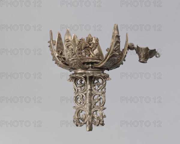 Lotus flower with Buddhist deities, 19th century, Nepal, cult object; bronze lotus mandala depicting lotus and Buddhist deities bodhisattva iconography, pairs of lovers, enlightenment; height 20.3 cm, width 8.0 cm., An ornate metallic object with a silvery-gray color is centered against a plain light gray background; the object features a decorative cup-like top with pointed petal-shaped extensions, detailed relief figures and floral motifs, a small ornate spout-like extension on one side, and an openwork cylindrical stem composed of intertwined scrolling patterns and leaf-like elements, all exhibiting a metallic sheen with darker gray shadows and subtle highlights.