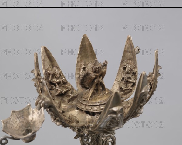 Lotus flower with Buddhist deities, 19th century, Nepal, cult object; bronze lotus mandala depicting Buddhist deities bodhisattva iconography, lotuses, pairs of lovers, enlightenment, height 20.3 cm, width 8.0 cm., The image shows a metallic sculptural object with a lotus-like arrangement of petal-shaped elements in a bronze or brass color, each petal containing small carved human or deity figures in a similar metallic hue; the central area features a raised circular platform with a more detailed figure, and surrounding the petals are ornate curved supports and decorative motifs in the same bronze/brass tones; the background is a smooth neutral gray.