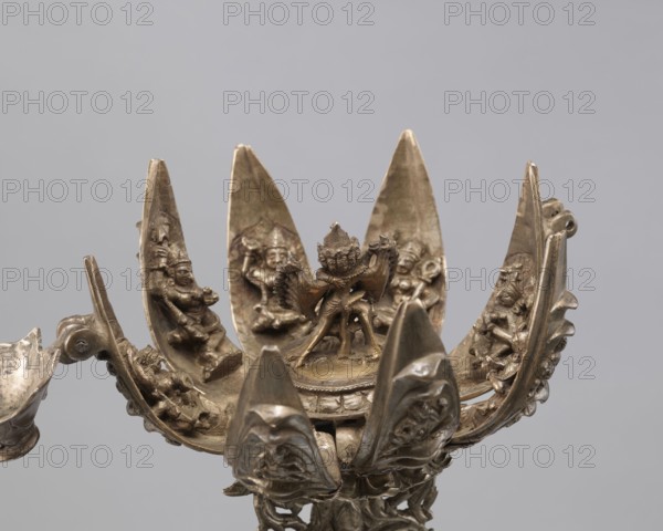 Lotus flower with Buddhist deities, 19th century, Nepal, cult object; bronze lotus mandala depicting Hevajra and Nairatmya with bodhisattva iconography, lotuses, pairs of lovers, symbolizing enlightenment; height 20.3 cm, width 8.0 cm., A metallic lotus-shaped object with a central sculpted figure seated on a raised platform surrounded by a group of similarly styled figures within petal-like panels; the petals curve upward and outward, each containing a small carved figure in relief, and the outer edges show ornate scrolling and textured details. The visible colors are bronze, brass, and muted gold tones with darker tarnished areas and subtle highlights. The background is a flat light gray.