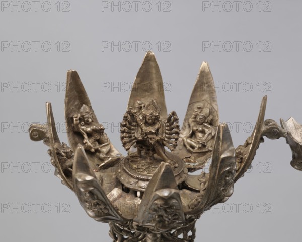 Lotus flower with Buddhist deities, 19th century, Nepal; bronze/lotus mandala depicting Hevajra and consort Nairatmya with bodhisattva iconography, pairs of lovers, lotus iconography; cult object; height 20.3 cm, width 8.0 cm; inscriptions: none specified; function: ritual/devotional., A metallic sculptural object with a central multi-armed figure seated on a raised circular platform, surrounded by a group of petal-like blades each bearing a seated figure in relief; the metal has a dark bronze to brown patina with areas of lighter, worn highlights and subtle greenish and reddish tones; the petals are pointed and arranged radially, creating an open flower-like form, with ornate curling supports and small decorative elements around the base and edges; the background is a smooth, neutral gray.