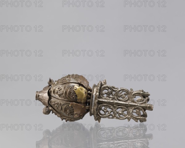 Lotus flower with Buddhist deities, 19th century, Nepal, bronze; cult object depicting lotus and Buddhist iconography lotuses, bodhisattva, pairs of lovers, enlightenment; height 20.3 cm, width 8.0 cm., An ornate metallic object rests on a reflective surface against a light gray background; the object shows a bulbous, rounded section with embossed floral and figurative reliefs and a cage-like, openwork extension with scrolling, vine-like patterns, all in shades of silver and brass with areas of darker patina and muted brownish tones, and the reflection of the object appears faintly on the surface below.