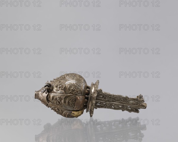 Lotus flower with Buddhist deities, 19th century, Nepal; bronze cult object depicting a lotus mandala with Buddhist deities bodhisattva imagery, pairs of lovers, iconography of lotus and enlightenment; height 20.3 cm, width 8.0 cm., An intricately carved metallic object lies horizontally against a pale gray background, showing a bulbous, ornate head with embossed floral and foliate motifs and a long, openwork handle with scrolling, lattice-like patterns; the piece appears mainly in shades of aged metallic tones including dull silver, bronze, and hints of darker gray, with small areas of warmer brass-colored highlights and subtle reflections of the object on the glossy surface beneath it.