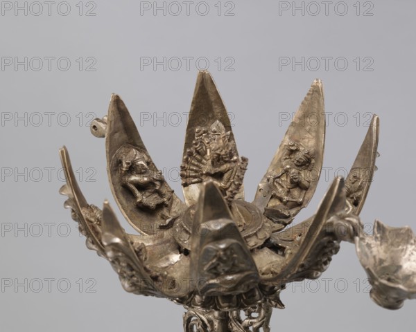 Lotus flower with Buddhist deities, 19th century, Nepal, lotus mandala of Hevajra and Nairatmya; Himalayan/Nepalese Buddhist art; cult object; bronze; height 20.3 cm, width 8.0 cm; iconography: lotus, pairs of lovers, bodhisattva, enlightenment., A metallic sculptural object with a patinated surface in shades of silver, grey, and bronze tones, shaped like an open lotus with several elongated petal-like elements radiating upward; each petal contains a small sculpted human or deity figure in relief, with visible limbs, headdresses, and ornamental details; the central area shows a raised circular base and additional intricate relief work, and the outer rim has sculpted decorative elements and curved forms; the background is a smooth, neutral pale grey.