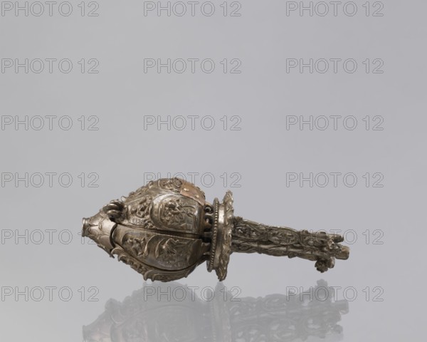 Lotus flower with Buddhist deities, 19th century, Nepal; lotus mandala cult object depicting Buddhist deities and iconography—lotuses, pairs of lovers, bodhisattva, enlightenment; bronze; height 20.3 cm, width 8.0 cm., A decorative metallic object rests horizontally against a light gray background, showing a carved bulbous end with ornate floral and leafy reliefs and a textured handle with intertwined vine-like patterns; the object and its reflection display colors of silver and dull gold with subtle brownish accents, and the scene features shades of light gray and muted off-white.