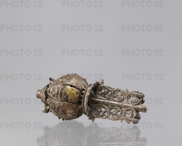 Lotus flower with Buddhist deities, 19th century, Nepal, lotus mandala depicting Hevajra and Nairatmya, Himalayan/Nepalese Buddhist art; cult object, bronze, height 20.3 cm, width 8.0 cm; iconography: lotus, pairs of lovers, bodhisattva, enlightenment., A decorative metal object rests horizontally on a reflective surface, showing a rounded bulbous end with embossed floral and leafy motifs and several applied plates, and a long openwork section composed of scrolling, intertwined vine-like elements; the metal appears in shades of silver and muted gold with areas of darker tarnish, and the background is a uniform pale gray.