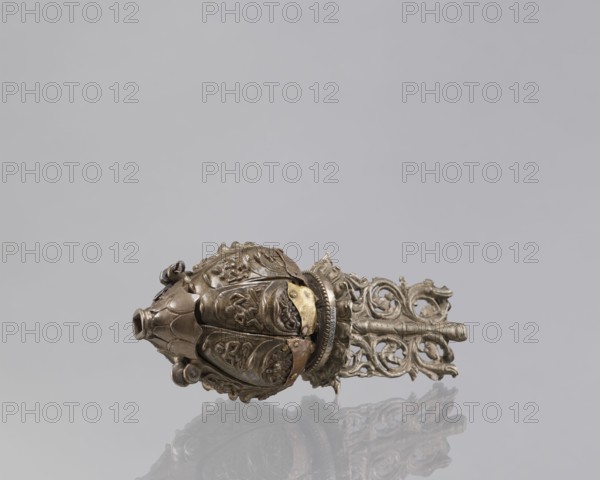 Lotus flower with Buddhist deities, 19th century, Nepal; bronze cult object in the form of a lotus mandala depicting Buddhist deities bodhisattva imagery, pairs of lovers, Hevajra/Nairatmya iconography, lotus iconography, associated with enlightenment; height 20.3 cm, width 8.0 cm., An ornate metallic object lies on a reflective surface against a plain light gray background, showing a bulbous, rounded head with petal-like segmented panels decorated with raised floral and foliate motifs and textured details, a short tubular opening at one end, and a complex openwork handle or extension with scrolling, pierced designs. The object displays metallic tones of aged bronze, brass, and silver with darker patina in crevices, and there is a subtle mirrored reflection of the piece on the surface below.