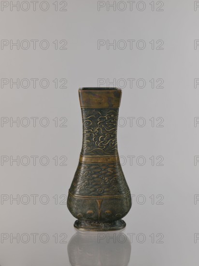 Hu-type vase, relief decoration, hu vessel, Chinese art, Chinese culture, meander ornament, ceremonial vessels, clouds, sea waves, ritual vessels, Xuande 1426-1435, Ming 1368-1644, Qing 1644-1911, backdated signatures, quadrilateral with rounded lower edges, with an inscription, 17th century, China, vessel, height 24.0 cm, width 12.0 cm, A single ornate vase stands against a plain light gray background, showing a tall narrow neck that widens into a rounded base with a small foot; the vase has a metallic appearance with dark bronze and muted gold tones, and its surface is covered in carved swirling patterns and wave-like motifs that repeat across the neck and body, with bands separating the sections and a subtle reflective sheen that creates darker and lighter areas on the metal.