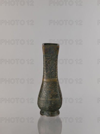 Hu-type vase, relief decoration, hu vessel, Chinese art, Chinese culture, meander ornament, ceremonial vessels, clouds, sea waves, ritual vessels, Xuande 1426–1435, Ming 1368–1644, Qing 1644–1911, backdated signatures, quadrilateral with rounded lower edges; with an inscription. 17th century, China; vessel; height 24.0 cm, width 12.0 cm., A single ornate vase stands upright against a neutral light gray background, showing a narrow rectangular mouth, a long tapering neck, and a rounded body that rests on a small circular base; the surface displays intricate raised patterns and swirling motifs, with visible wear and texture variations; the colors present include dark bronze, muted greenish patina, hints of brown and black, and subtle metallic highlights; the vase casts a soft reflection on the smooth surface beneath it.