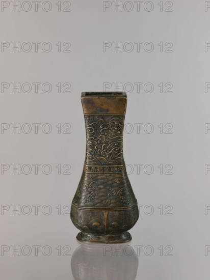 Hu-type vase, relief decoration; hu vessel; Chinese art; Chinese culture; meander ornament; ceremonial and ritual vessel with clouds and sea waves motifs; Xuande 1426–1435, Ming 1368–1644, Qing 1644–1911; backdated signatures; quadrilateral with rounded lower edges; with an inscription; 17th century ; China; height 24.0 cm, width 12.0 cm., An ornate metal vessel stands upright against a neutral gray background, its surface showing a patina of dark bronze and lighter brass tones; the top section is rectangular and slightly weathered with brownish and golden hues, the neck below it decorated with intricate swirling wave-like engravings in darker and lighter bronze, the midsection broadens into a bulbous body covered in carved patterns resembling stylized clouds and flowing waves with subtle contrast between dark and warm metallic shades, a band separates this from the lower rounded base which features repeated geometric and curvilinear motifs in similar bronze and brass colors, the piece...