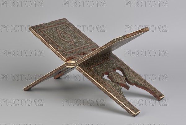 Quran lectern rahla, Islamic art, Persian art, Qajar dynasty 1785–1925, geometric ornament, stands, rahla, Quran, Persian culture, 19th century, Iran, piece of furniture; unfolded: length 21.5 cm, width 41.4 cm; folded: length 41.5 cm, width 16.5 cm., An ornate folding wooden stand displayed open in an X shape, showing carved and inlaid panels with dense small floral or geometric patterns in dark green and brown, bordered by narrow bands of red and cream, with visible wood grain in reddish-brown on the structural edges and supports, set against a plain light gray background.