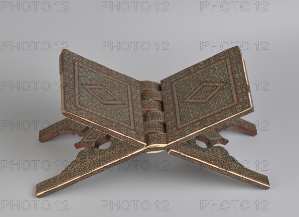 Quran lectern ra?la, maker, 19th century Qajar dynasty, Iran; Persian Islamic art; geometric ornament; piece of furniture; function: stands/ra?la for the Quran; unfolded: length 21.5 cm, width 41.4 cm; folded: length 41.5 cm, width 16.5 cm., An open wooden book stand with two hinged panels rests on a pale gray surface against a light gray background; the stand is decorated all over with intricate repeating geometric and floral patterns in dark brown, gold, red, black, and green, with lighter cream-colored edging along the outer borders, and the carved legs form intersecting supports with matching ornate designs.
