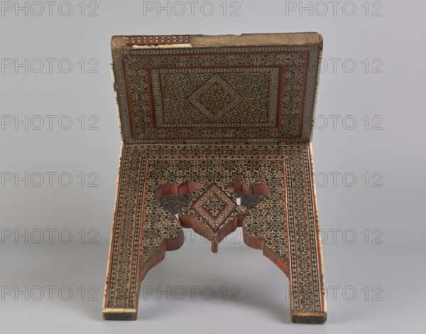 Quran lectern rahla, Islamic art, Persian art, Qajar dynasty 1785–1925, geometric ornament, stands, rahla, Quran, Persian culture, 19th century, Iran, piece of furniture; unfolded: length 21.5 cm, width 41.4 cm; folded: length 41.5 cm, width 16.5 cm., An ornate folding wooden stand with intricate geometric inlaid patterns covering its surfaces, showing a rectangular top panel hinged to an angled lower panel that splits into two carved legs; the patterning forms concentric framed designs with a central diamond motif on both panels, and the lower panel includes a decorative cutout where the legs meet; visible colors include brown from the wood, dark brown or black in the inlay, red accents, cream or beige highlights, and small touches of green, all set against a neutral light gray background.