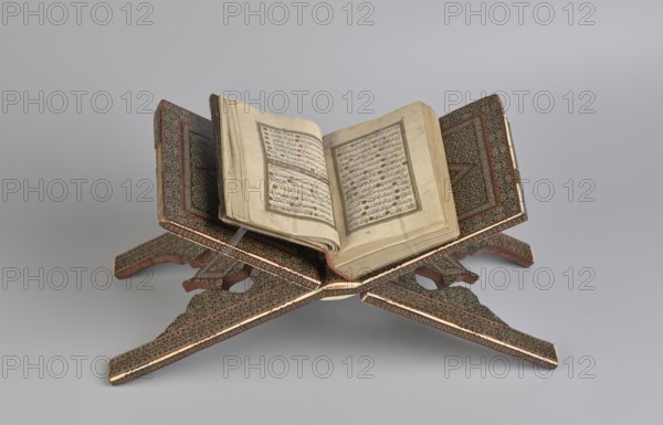 Quran lectern rahla, Islamic art, Persian art, Qajar dynasty 1785–1925, geometric ornament, stands, rahla, Quran, Persian culture, 19th century, Iran, piece of furniture; unfolded: length 21.5 cm, width 41.4 cm; folded: length 41.5 cm, width 16.5 cm., An open book with aged, off-white pages rests on an ornately carved wooden stand with an X-shaped support; the stand shows detailed geometric and floral patterns in dark brown, reddish-brown, and cream with accents of green and gold; the book's pages are cream with visible lines of script and decorative elements in muted brown and faded black; the background is a smooth light gray.