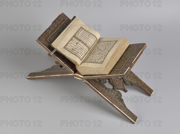 Quran lectern rahla, Islamic art, Persian art, Qajar dynasty 1785–1925, geometric ornament, stands, rahla, Quran, Persian culture, 19th century, Iran, piece of furniture; unfolded: length 21.5 cm, width 41.4 cm; folded: length 41.5 cm, width 16.5 cm., An open book rests on an ornate folding stand with carved interlocking panels; the book's pages are creamy beige with darker edges and the stand displays intricate patterned decoration in colors including brown, dark red, gold, black, and cream, with areas of wood grain visible; the background is a smooth gradient of light gray.