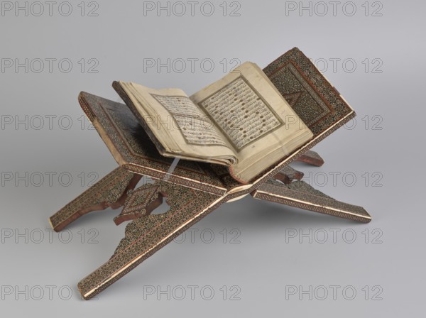 Quran lectern rahla, 19th century, Qajar dynasty 1785–1925, Iran; Persian; Islamic art; geometric ornament; stands; piece of furniture; function: lectern for the Quran; unfolded: length 21.5 cm, width 41.4 cm; folded: length 41.5 cm, width 16.5 cm., A decorative folding wooden book stand painted with intricate repeating geometric and floral patterns in brown, dark green, red, white, and gold holds an open aged book with cream-colored pages; the stand has a pair of crossed slotted panels with carved supports and visible edges showing red and white bordering, and the background is a smooth light gray.
