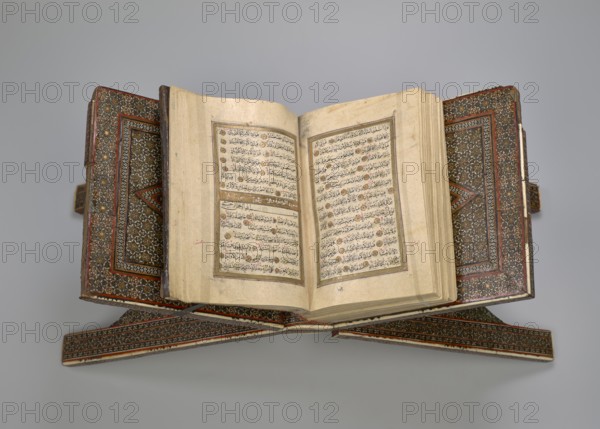 Quran lectern rahla, Islamic art, Persian art, Qajar dynasty 1785–1925, geometric ornament, stands, rahla, Quran, Persian culture, 19th century, Iran, piece of furniture; unfolded: length 21.5 cm, width 41.4 cm; folded: length 41.5 cm, width 16.5 cm, An open ornate book rests on an intricately decorated wooden stand, showing two facing pages with dense lines of dark script surrounded by rectangular decorative borders; the book cover and stand feature repeating geometric and floral patterns in shades of brown, red, gold, black, and white, while the pages are aged beige with some darker spots and light brown edges, all set against a smooth light gray background.