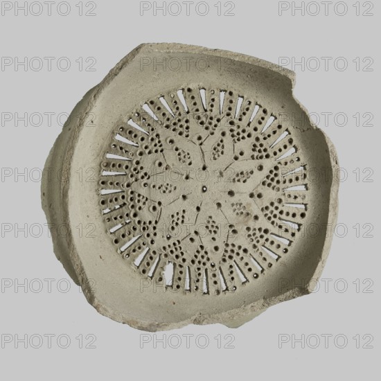 Strainer — jug filter;, Islamic art, Arabic; Mamluk Sultanate 1250–1517; Arabic/Egyptian ceramics; star ornament; everyday life in the Middle East; 13th–15th century, Egypt; vessel; height 4.5 cm, width 11 cm, diameter 10.3 cm., A circular, shallow object with a scalloped rim sits against a plain light gray background; the object is a muted beige-gray color with a rough, stone-like texture and irregular edges, and its central area features an intricate radial pattern of small round perforations and elongated slits forming floral and geometric motifs, creating a lace-like appearance with shadowed holes and a few tiny darker specks on the surface.