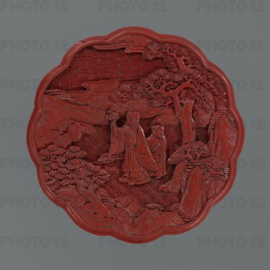 Box with a lid, Chinese, Qing 1644–1911, 2nd half of the 18th century. Lacquered box decorated with figurative scenes of sages and scholars with servants in landscape settings pine trees, rocks, rosette and geometric ornament, kikko pattern. Vessel; height 11.0 cm, width 4.8 cm., A carved circular plaque with a scalloped edge, predominantly deep red, set against a neutral gray background; the plaque shows a high-relief scene with two robed human figures standing amid stylized natural elements including a large textured tree, layered rock formations, and patterned ground; intricate carved foliage and fan-shaped leaves surround the figures, and the background includes a woven or crosshatched surface texture; the carving displays varied surface depths and fine linear details throughout, all rendered in shades of red with the surrounding backdrop in gray.