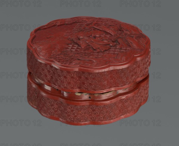 Box with a lid, Chinese, Qing 1644–1911, 2nd half of the 18th century. Lacquered vessel decorated with figurative scenes of sages, scholars, and servants amid landscape motifs pine trees, rocks, rosettes, kikko tortoiseshell and geometric ornament. Height 11.0 cm, width 4.8 cm., A round, scalloped-edge carved box sits against a plain gray background; the box is predominantly deep red with carved surface textures showing raised patterns and relief scenes on the lid and sides, and there are small areas where the red finish is chipped revealing a few patches of darker and lighter tones beneath.