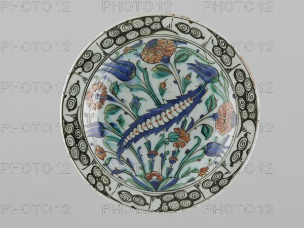 Plate, Islamic art, Turkish, Ottoman dynasty 1299–1922, floral motifs including tulips, carnations, saz, early 17th century, Iznik, Turkey; vessel; height 29.0 cm, width 5.7 cm., A round ceramic plate with a decorative floral design fills the frame, showing a central motif of stylized flowers and leaves in shades of blue, green, and orange on a white background, with a prominent curved blue element edged with a white bead-like pattern. The rim features a repeating black spiral pattern on white, and the inner field contains several tulip-like shapes, rounded blossoms, and slender stems with leaves in green and turquoise, plus small orange flower accents. The overall colors visible are white, blue, green, black, orange, and a touch of turquoise.