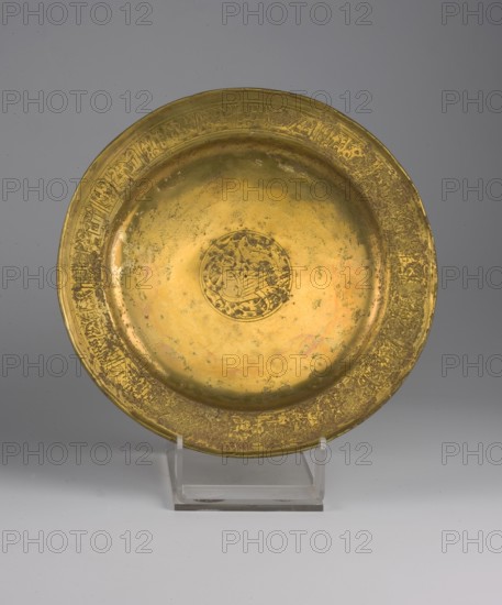 Flat metal plate with a slightly raised rim, 13th century, Iran. Diameter 20.8 cm, height 2.2 cm, weight 0.22 kg. The rim bears a band of Arabic inscription in kufic script reading names and blessings such as omnipotence, divine majesty, splendor, fortune... blessings, well-being, prosperity, grace. Some letters are abraded by corrosion and two words remain undeciphered; the words bahja splendor, magnificence and subha majesty are the most plausible readings. The central field depicts a harpy — a bird with a human feminine head, with wavy short hair styled in court fashion and a halo around the raised head. In pre-Islamic and later belief the harpy guarded the divine other world against human intrusion; combined with the benedictory inscription, the image likely conveyed the owner's hope for eternal life., A round metallic plate with a golden color and areas of darker patina sits upright on a clear plastic stand against a smooth gray background that fades to a lighter gray at the botto...
