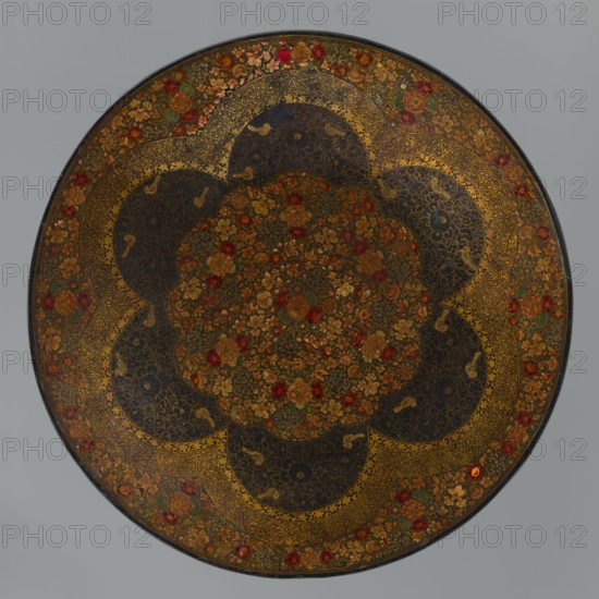 Plate, Persian, Qajar dynasty 1785–1925, 19th century, Iran; ceramic vessel with floral ornament flowers; height 7.5 cm, diameter 42.0 cm., A circular decorative object with an ornate floral pattern, featuring a central area densely filled with a group of painted flowers in red, gold, green, and brown tones, surrounded by a scalloped band of dark panels with intricate gold filigree and small bird motifs, then an outer ring decorated with a repeating floral border in shades of red, gold, green, and brown against a dark background, all set on a neutral gray backdrop.