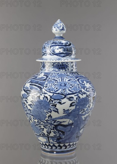 Vase with cover, unknown ceramist, Japanese, Edo period Genroku–Kyoho, ca. 1690–1730, Arita porcelain, blue-and-white; decorated with phoenixes, hares/rabbits, peonies, chrysanthemums, and lotuses; one of the four elements of a fireplace set; height 59.0 cm, diameter 33.0 cm., A lidded porcelain vase standing against a plain background, decorated in blue and white. The vessel has a bulbous body tapering to a narrower base and a matching lid topped with a pointed finial. The surface shows dense painted ornamentation including floral motifs, leafy branches, stylized panels, and a bird in flight among the foliage. The blue decoration varies in tone from deep indigo to lighter blue washes, set against a white ground. The lid and neck carry coordinated patterned bands and small decorative panels, and the base has a repeating border motif. The background color is a neutral gray.
