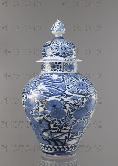 Vase with cover, unknown ceramist, Japanese, Edo period Genroku–Kyoho, ca. 1690–1730, Arita porcelain, blue-and-white porcelain, decorated with phoenixes, hares/rabbits, peonies, chrysanthemums, lotuses; one of the four elements of a fireplace set; vessel; height 59.0 cm, diameter 33.0 cm., A lidded ceramic vase with a bulbous body and a pointed finial lid decorated in blue and white, featuring floral and foliate motifs and stylized patterns across its surface; the vase shows glossy highlights and a reflective base, and the background is a uniform gray.