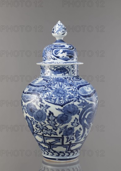 Vase with cover, unknown ceramist, Japanese, Edo period Genroku–Kyoho, ca. 1690–1730, Arita; porcelain, blue-and-white; decorated with phoenixes, hares/rabbits, peonies, chrysanthemums, lotuses — kachoga flowers-and-birds motif; one of a set of four; vessel height 58.0 cm, diameter 35.5 cm., A lidded ceramic vase displayed against a plain gray background, predominantly white with intricate blue painted decoration covering its surface; the lid has a pointed finial with blue floral and scroll motifs, the rounded shoulder and body feature detailed blue floral patterns, leafy stems, and decorative panels, with additional scrolling and geometric banding near the base and rim; the overall color palette is blue and white with subtle reflections on the glazed surface.