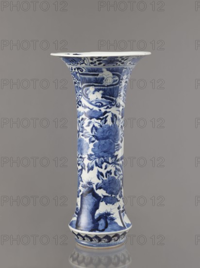 Vase, Japanese, Edo 1603–1868, Genroku 1688–1704–Kyoho 1716–1736, ca. 1690–1730, blue-and-white porcelain; Arita; vessel; height 39.0 cm, diameter 20.3 cm. Decoration: hares/rabbits, peonies, lotuses, phoenix hoo, kacho/kachôga flowers and birds. One of four elements of a set SKAZsz 314/1–4., A tall flared ceramic vase stands against a neutral gray background, decorated in a blue-and-white palette; the surface shows floral motifs and leafy patterns in varying shades of blue on a white ground, with decorative bands near the rim and base featuring stylized wave and petal designs, and areas depicting rounded rock-like forms and curved shapes among the flowers.