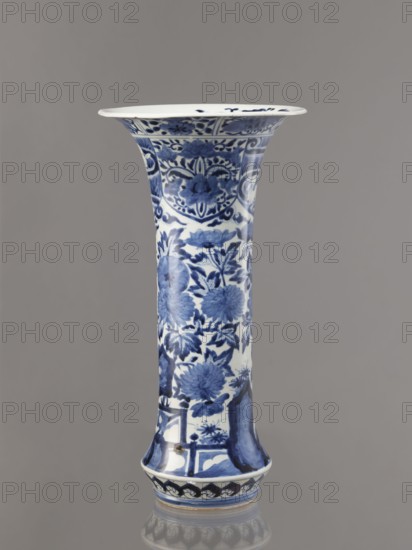 Vase, Japanese, Edo period 1603–1868, Genroku–Kyoho ca. 1690–1730. Blue-and-white porcelain from Arita, decorated with hares/rabbits, peonies, lotuses, and phoenixes kacho/kachôga motifs. One of four elements of a set; vessel height 39.0 cm, diameter 20.3 cm., A tall, flared ceramic vase with a glossy white base decorated in blue floral and foliate patterns, featuring large stylized flowers and leaves arranged vertically along its narrow body, a flared rim with decorative bands and motifs, and a slightly flared foot with a repeating decorative border; the background is a smooth, neutral gray, and the vase shows shades of deep blue and lighter blue on white with subtle reflections on its surface.