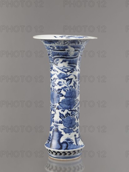 Vase, Japanese, Edo period 1603–1868, Genroku–Kyoho period ca. 1690–1730, blue-and-white porcelain decorated with phoenixes, hares/rabbits, peonies, chrysanthemums, lotuses—kacho flowers and birds / kachôga; one of four elements of a set, Arita, vessel, height 39.0 cm, diameter 20.3 cm, A tall, slender ceramic vase with a flared rim, predominantly white with blue painted decoration, showing floral motifs and stylized foliage along its elongated body, a band of decorative patterns near the top and a repeating wave-like motif around the base; the blue ranges from deep indigo to lighter cobalt tones, and the background is white, set against a neutral gray backdrop.