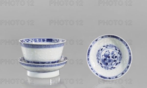 Bowl on a base with a lid, ceramics, rice pattern, geometric ornament, plant ornament, Chinese art/culture, rice porcelain, Qing 1644–1911, late 18th century, China, Floral arrangement in a circle at the bottom. Signed. Vessel, height 10 cm., A group of ceramic bowls with blue and white coloring sits against a gray background; one stack of bowls is positioned on the left showing the exterior with blue patterned rims and a white body, while a single bowl on the right is shown face-up revealing a blue floral design in the center and a decorative blue border around the inner rim; reflections of the bowls appear faintly on the surface below.