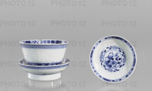 Bowl on a base with a lid;, Ceramic rice porcelain, Qing 1644–1911, late 18th century, China. Geometric ornament with rice pattern and plant/Chinese floral motifs; floral arrangement in a circle at the bottom. Signed. Vessel, height 10 cm., A group of ceramic bowls displayed against a neutral gray background, with a few stacked on the left and one shown upright on the right; the ceramics are glossy white with blue decorative patterns along rims and interiors, featuring floral motifs and geometric borders; the left stack shows a chip on the rim of the top bowl and reflections on the surface beneath, while the right bowl displays a circular floral design in its center and a patterned rim; visible colors include white, blue, and gray.