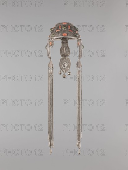 Headgear ornament coif, 19th century, Mongolia. Women's headgear / head ornament made of silver with ear cuffs; filigree. Height 57.0 cm, diameter 14.0 cm., An ornate metallic headdress with a domed top decorated with oval and rounded cabochon stones in shades of coral and green, featuring intricate filigree and textured silverwork; two curved side pieces extend downward from the dome and connect to a central vertical pendant composed of linked engraved segments and small spherical bells, with long delicate silver chains hanging from each side ending in small cylindrical metal cones; the colors visible are silver, coral, green, and subtle variations of gray against a neutral light-gray background.