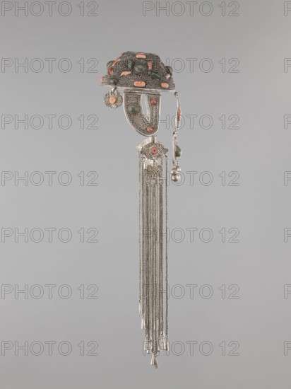 Headgear ornament coif, coif; jewelry; Mongolian artistic crafts; headgear and head ornaments for women. Made of silver with ear cuffs; filigree. 19th century, Mongolia. Clothing accessory. Height 57.0 cm; diameter 14.0 cm., A decorative metal pendant with an ornate upper section featuring textured silver surfaces and embedded green and coral-colored stones, a few filigree elements and small dangling ornaments, a curved silver plate with green and coral-colored round stones attached below, a floral filigree connector with a coral-colored center and several long silver chains hanging down ending in small cone-shaped metal beads; visible colors include silver, green, coral, and a neutral gray background.