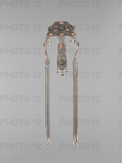 Headgear ornament coif, coif, jewelry, Mongolian art, artistic crafts, headgear, head ornaments, women's ornaments. Made of silver with ear cuffs; filigree. 19th century, Mongolia. Clothing accessory. Height 57.0 cm, diameter 14.0 cm., An ornate metallic headdress piece hangs against a plain light gray background, featuring an arched top section decorated with rounded colored stones in shades of coral and green set into textured silver metal; dangling from the top are three linked filigree panels with an oval green stone in the central lower panel and small spherical silver beads suspended beneath it; two long groups of fine silver chains extend downward from either side, each ending in small tapered metallic tassels, and there are small coral-colored accents and green beads visible on the side elements, with overall colors including silver, coral, green, and the neutral gray of the background.