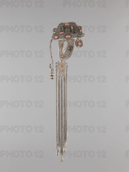 Headgear ornament coif, 19th century, Mongolia. Made of silver with ear cuffs; filigree. Category: coif/headgear/head ornament for women; clothing accessory. Height 57.0 cm; diameter 14.0 cm., An ornamental metal hairpiece with intricate filigree and textured surfaces decorated with a few oval and round cabochon stones in green and coral colors, featuring a domed top section with beaded edging, a curved plate element beneath, a small dangling decorative charm and a cluster of two small spherical pendants, and a group of long fine metal chains ending in tapered metal cones, all shown against a plain light gray background.