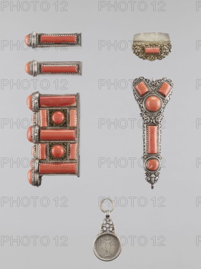 Braid ornament, braid ornament jewelry, artistic craft; braid ornament for women's dress, 19th–20th century, Mongolia; materials: jewelry; height 3.0 cm, width 7.8 cm., A vertical arrangement of several ornamental metal pieces set against a pale gray background, featuring a group of jewelry-like objects with coral-colored stones framed in silver-toned metal, including a rectangular cluster with rounded coral cabochons and long coral bars, a trio of slim rectangular pieces each capped with a rounded coral end, an ornate pendant-like piece with a heart-shaped top and vertical coral panels, a small scalloped metal element with a coral rectangle, and a round coin-like medallion suspended from a decorative silver loop; visible colors include coral, silver, and pale gray.