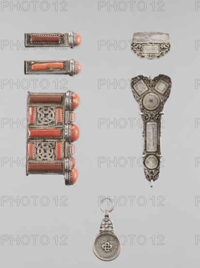 Braid ornament, braid ornament jewelry, artistic crafts, Mongolian art, ornaments, braids, women's ornaments, 19th–20th century, Mongolia; materials: jewelry; dimensions: height 3.0 cm, width 7.8 cm., A vertical arrangement of several metallic jewelry pieces on a pale gray background, including a rectangular decorative cuff with intricate openwork panels and coral-colored oval cabochons set along one side, two small matching rectangular pieces with coral-colored insets and rounded coral-tipped ends, an elongated ornate metal pendant with filigree edges and circular recessed areas, a small scalloped metal plate with decorative cutwork, and a round metal medallion charm with a linked lattice loop and a raised knot motif; visible colors include silver, coral, brown, and pale gray.