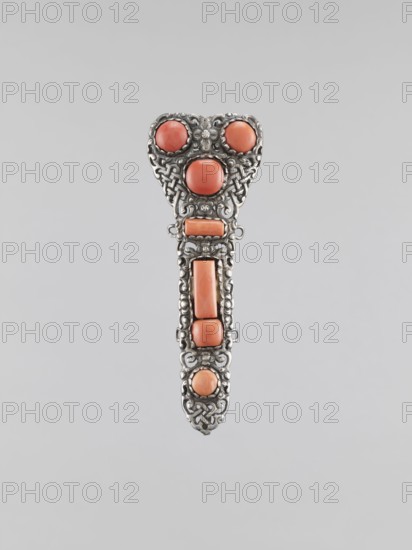 Clasp buckle, clasp, jewelry, Mongolian art, artistic crafts, braid ornament, women's jewelry. The clasp consists of two longitudinal elements and one transverse element, decorated with 17 pieces of coral. Late 18th century, Mongolia. Height 10 cm., A vertically oriented ornamental object with an elongated teardrop shape featuring a filigree metal framework in a silvery tone and set with several rounded and rectangular coral-colored stones, the top portion forming a broader heart-like outline with three round coral-colored cabochons surrounded by intricate silver scrollwork, a central elongated section with rectangular coral-colored pieces framed by interlaced silver patterns, and a narrower pointed lower end with a final round coral-colored stone encased in decorative silverwork; the background is a plain light gray.