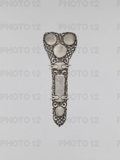Clasp buckle, clasp, jewelry, Mongolian art, artistic crafts, braid ornaments, women's jewelry. The clasp consists of two longitudinal elements and one transverse element. Decorated with 17 pieces of coral. Late 18th century, Mongolia. Height 10 cm., A decorative metallic object with an elongated, ornate shape suspended against a plain light gray background. The object is primarily silver in color with darker gray areas where shadows and patina appear. The top section is wider with a rounded triangular outline and features three circular raised discs surrounded by intricate openwork filigree. The central shaft narrows and displays a rectangular recessed panel flanked by small looped fasteners and more scroll-like openwork. The lower end tapers into a pointed form with another circular raised disc set within continued filigree and small hooked elements. The surface shows reflective highlights and subtle variations of silver and gray tones.