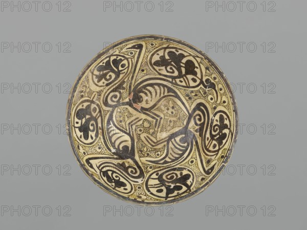 Bowl, maker, 10th century; Nishapur, Iran; Persian Islamic ceramic vessel decorated with images of birds and palmette ornament; height 12.1 cm; diameter 26.0 cm; weight 1.11 kg., A round ceramic bowl viewed from above with a decorative interior showing swirling organic patterns and stylized animal-like figures in black on a yellowish-cream background, with areas of darker brown and small speckled dotting filling spaces; the rim shows a thin dark band and some worn spots, and the whole piece sits against a flat medium-gray backdrop.
