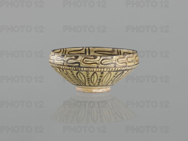 Bowl, 10th century, Nishapur, Iran; Persian Islamic ceramic vessel decorated with images of birds and palmettes; weight 1.11 kg, height 12.1 cm, diameter 26.0 cm., A shallow bowl with a rounded body and small foot, decorated all around with curving and leaflike painted motifs; the bowl shows yellow, black, brown, beige, and a hint of reddish tones, and it sits on a smooth gray surface that produces a soft reflection of the bowl.