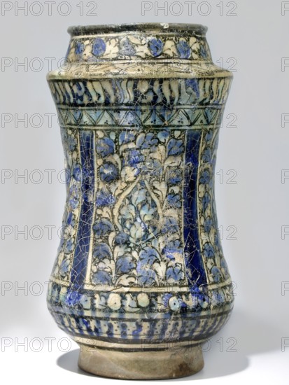 Albarello, 14th century Iran, Persian, Islamic art. Ceramic albarello vessel with floral/plant motifs, weight 0.99 kg, height 25 cm, diameter 13.5 cm., A tall ceramic vessel with a slightly flared rim, a bulging midsection, and a narrower base, decorated all over with an intricate painted pattern of floral and vine motifs; the surface shows a network of fine cracks. The visible colors include shades of blue ranging from deep cobalt to lighter sky blue, off-white or cream, black outlining, and areas of beige or light brown on the unglazed base. Several vertical panels of dense floral decoration alternate with darker blue vertical bands, and there is a band of repeating floral motifs near the top and another decorative band near the bottom. The vessel sits on a short, unglazed foot and casts a soft shadow on the surface beneath it.