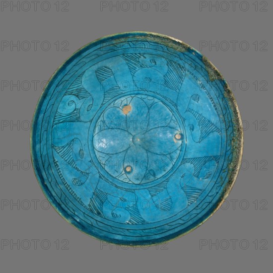 Bowl, Islamic art, Persian culture, ceramics, Persian ceramics, inscriptions, geometric motifs, 12th–13th century, Afghanistan; Iran place of origin, vessel, weight 0.344 kg, height 8.2 cm, diameter 19.5 cm, A circular ceramic plate viewed from above with a vivid turquoise blue glaze, showing a central floral motif with petal shapes and a few small worn spots revealing a tan underlayer, surrounded by a ring of flowing abstract curvilinear patterns outlined in darker blue or black, with additional fine hatch marks and line work within the decorative bands; the rim shows areas of darker greenish and brown discoloration and minor roughness, and the background behind the plate is a uniform medium gray.